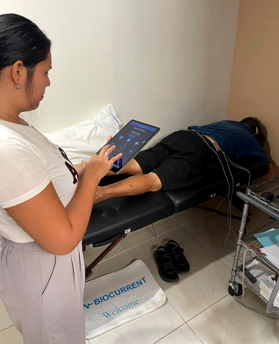 A Biocurrent clinician runs a 30-minute session from the tablet app while a study participant lies prone with surface electrodes placed along the spine, at the trial clinic in Puerto Vallarta, Mexico.